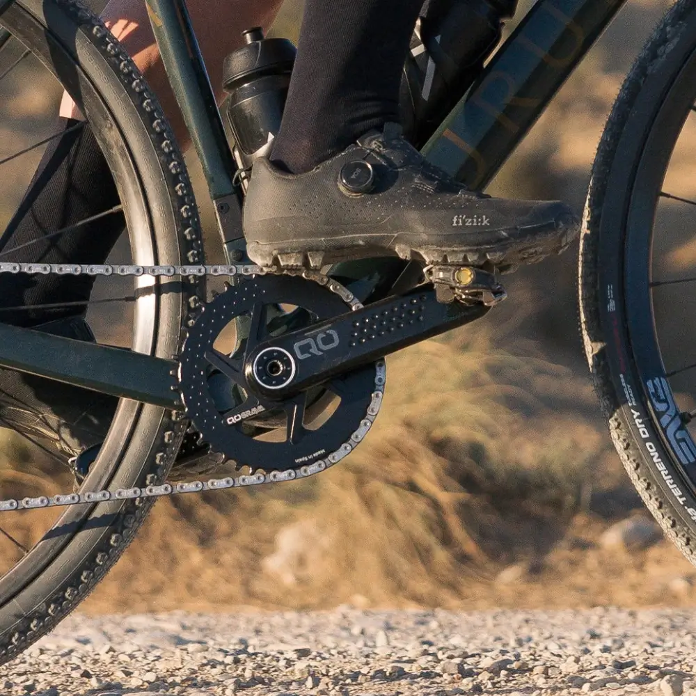 A black QO GRAVA Gravel Crankset installed on a dark green gravel bike with a rider clipped in using black cycling shoes.