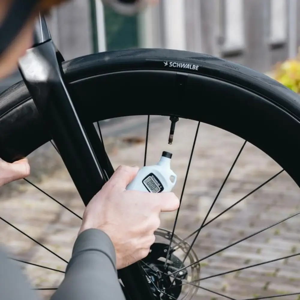 A white Schwalbe Airmax Pro Digital Tire Pressure Gauge being used on a black road bike to check a tire's pressure.